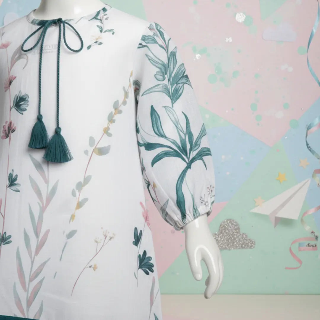 Close up of girls white tunic featuring teal botanical print, tassel tie-up neckline and ribbon hem details