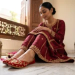 Pakistani woman in maroon gold embroidered suit seated on marble floor near carved wooden window wearing red velvet zardozi khussa jutti — handcrafted jutti online Pakistan — Applique Threads